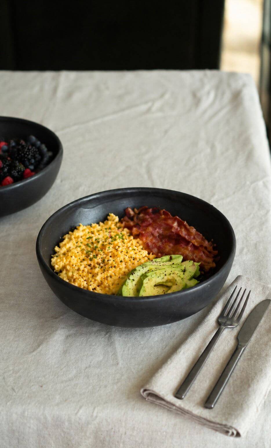 Black Matte Stoneware Pasta Bowl filled with scrambled eggs, bacon, and avocado on a dining table.