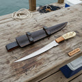 Handmade fillet knife with engraved handle beside leather sheath on wooden surface, designed for precise fish preparation.