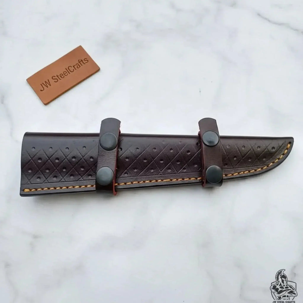 Leather sheath for handmade fillet knife with engraved handle, secured with snap buttons, showcased on marble surface.