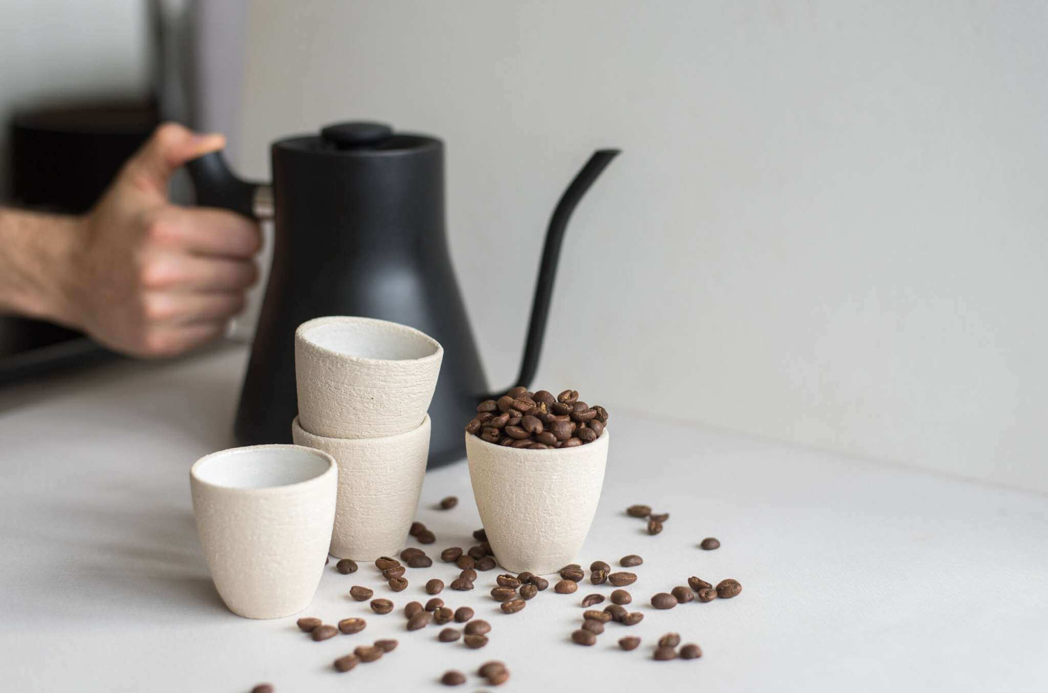 Handmade stoneware espresso cup and coffee beans with a black kettle in a cozy setup.