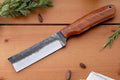 Bull Cutter Field Knife on wooden surface with greenery and spices, highlighting its custom design and build quality.